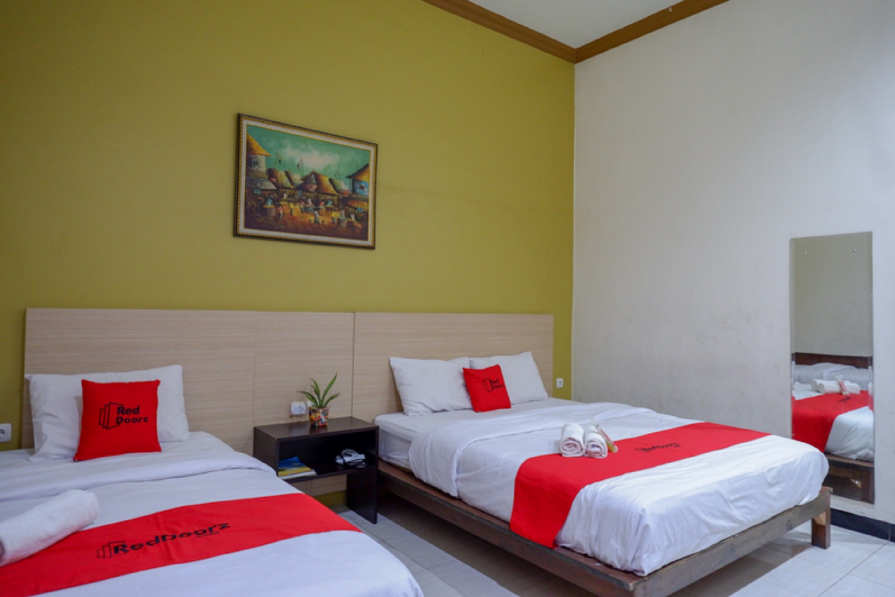 A hotel room with two beds, each with a red and white bedspread, a painting on the wall, and a nightstand with a plant.