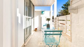 Un estrecho pasillo exterior con una pared blanca, una ventana con rejas, una mesa pequeña con una planta en maceta y una silla azul.