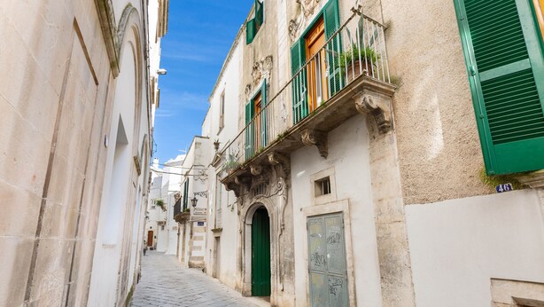 A narrow cobblestone street flanked by historic buildings with green shutters and ornate balconies. - 2163 Palazzo Grassi (Martina Franca)