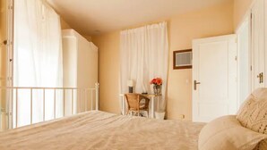 A bedroom with a bed, a chair, a small table with a vase of flowers, and a door. - Alohamundi San Diego (Sevilla)
