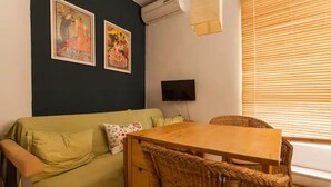 A room with a green sofa, a wooden table, wicker chairs, two posters on the wall, a flat-screen TV, and a window with blinds. - Alohamundi San Diego (Sevilla)