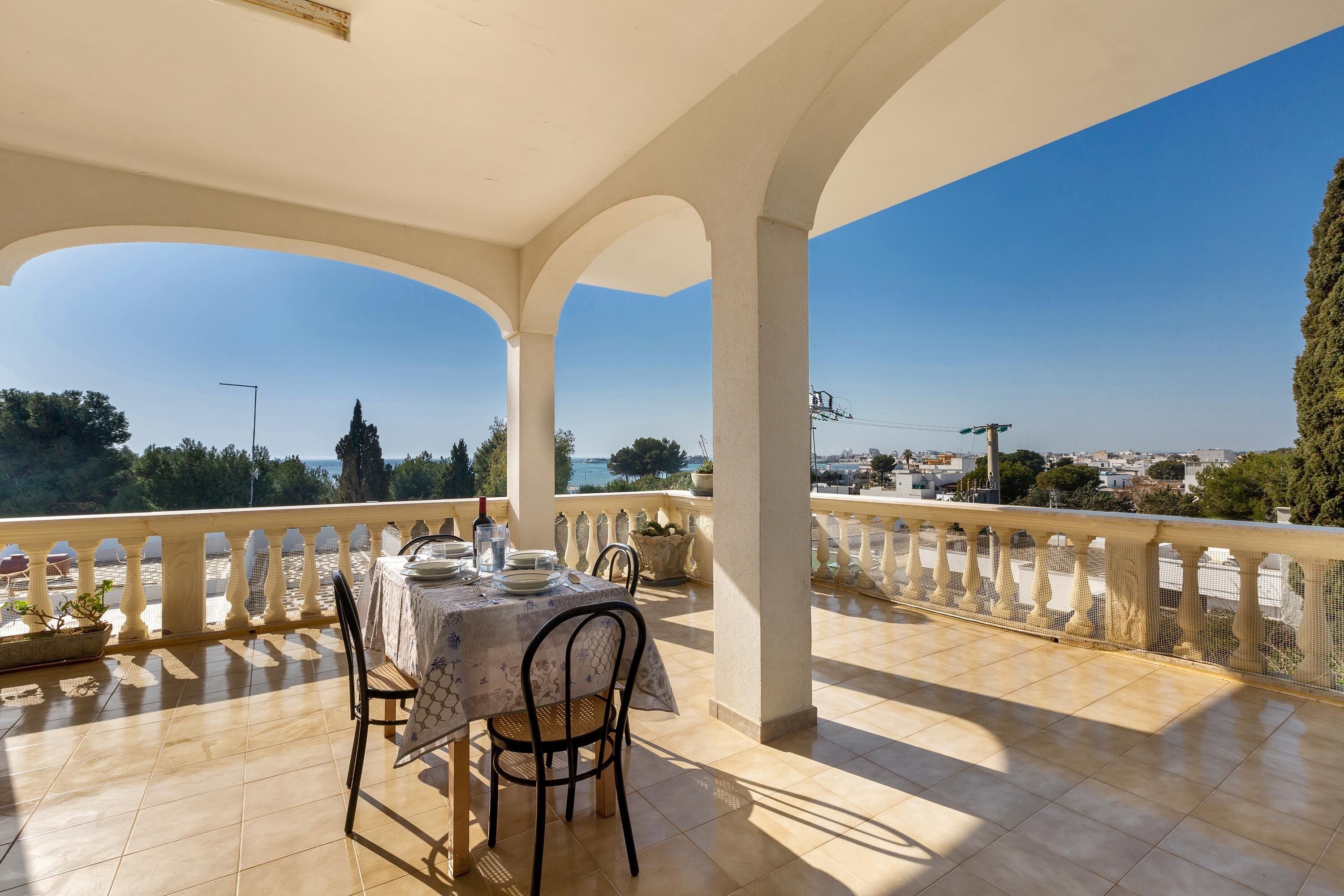 A covered outdoor dining area with a table set for two, overlooking a scenic view of the sea and cityscape.