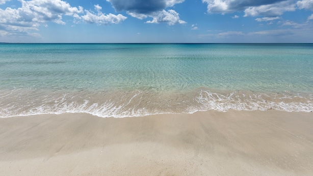 Une plage à l’eau turquoise et limpide, bordée d’un rivage sablonneux, sous un ciel partiellement nuageux.