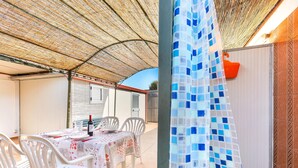 Une terrasse extérieure avec une table dressée pour le repas, des chaises blanches et un espace abrité doté d’un plafond en osier.