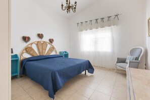 A bedroom with a bed, a chair, a nightstand, a dresser, and a chandelier.