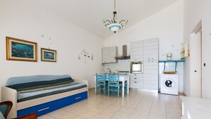 A compact living space with a sofa, a dining table, chairs, a kitchen area with a washing machine, and a ceiling light fixture. - 2299 Villetta Noemi (Porto Cesareo)