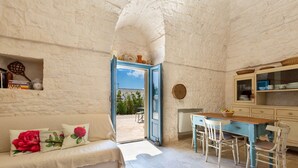 Un interior rústico con una pared de piedra, un sofá blanco con cojines florales rojos, una mesa de comedor de madera y una puerta azul que da al exterior.
