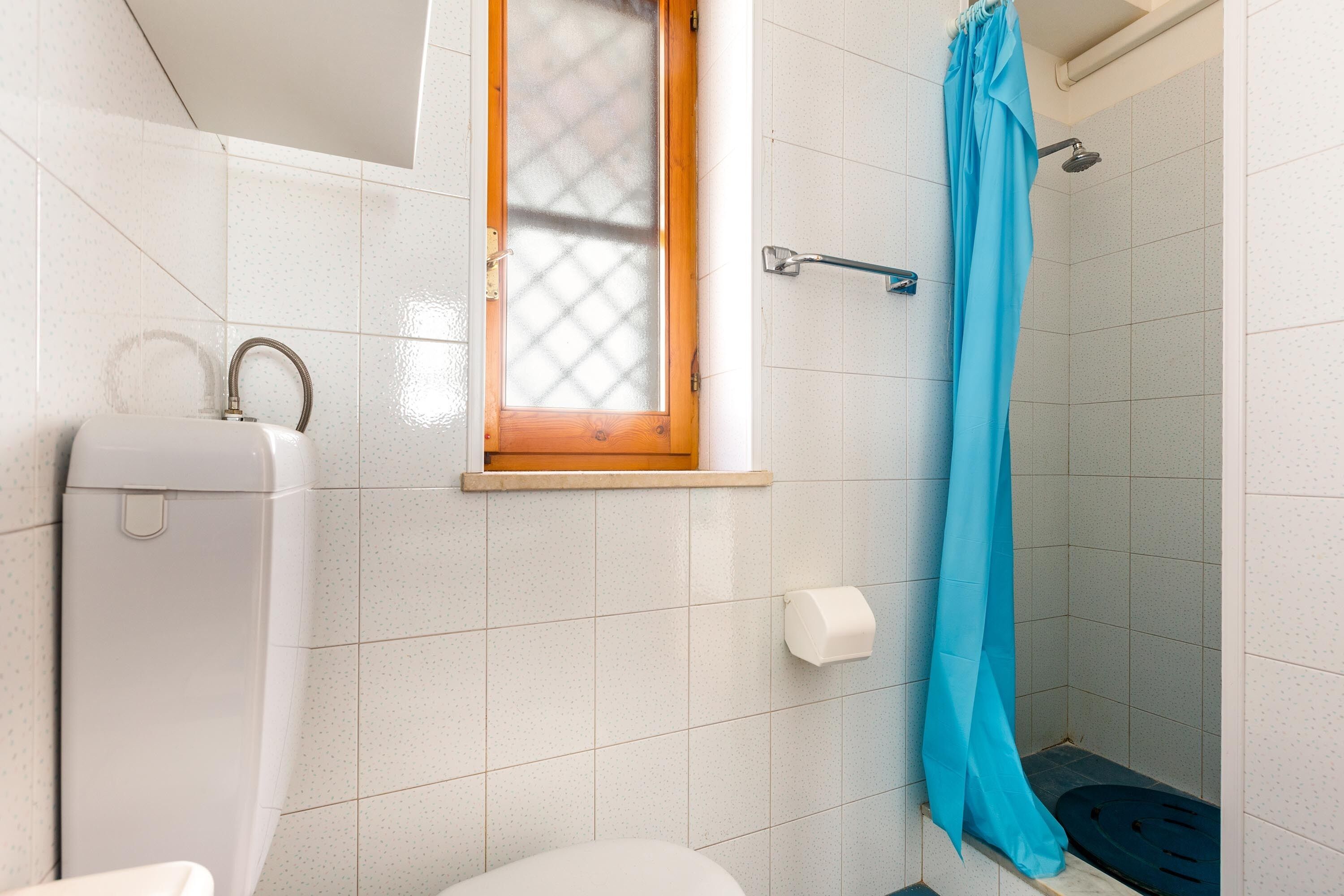 A bathroom with a toilet, a shower with a blue curtain, and a window with a wooden frame.