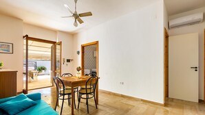 A room with a wooden dining table, chairs, a blue sofa, a ceiling fan, and an air conditioning unit. - 2019 Villetta del Colle (Porto Cesareo)