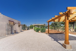A driveway leading to a building with a gated entrance, a pergola structure, and a parking area with a clear sky.