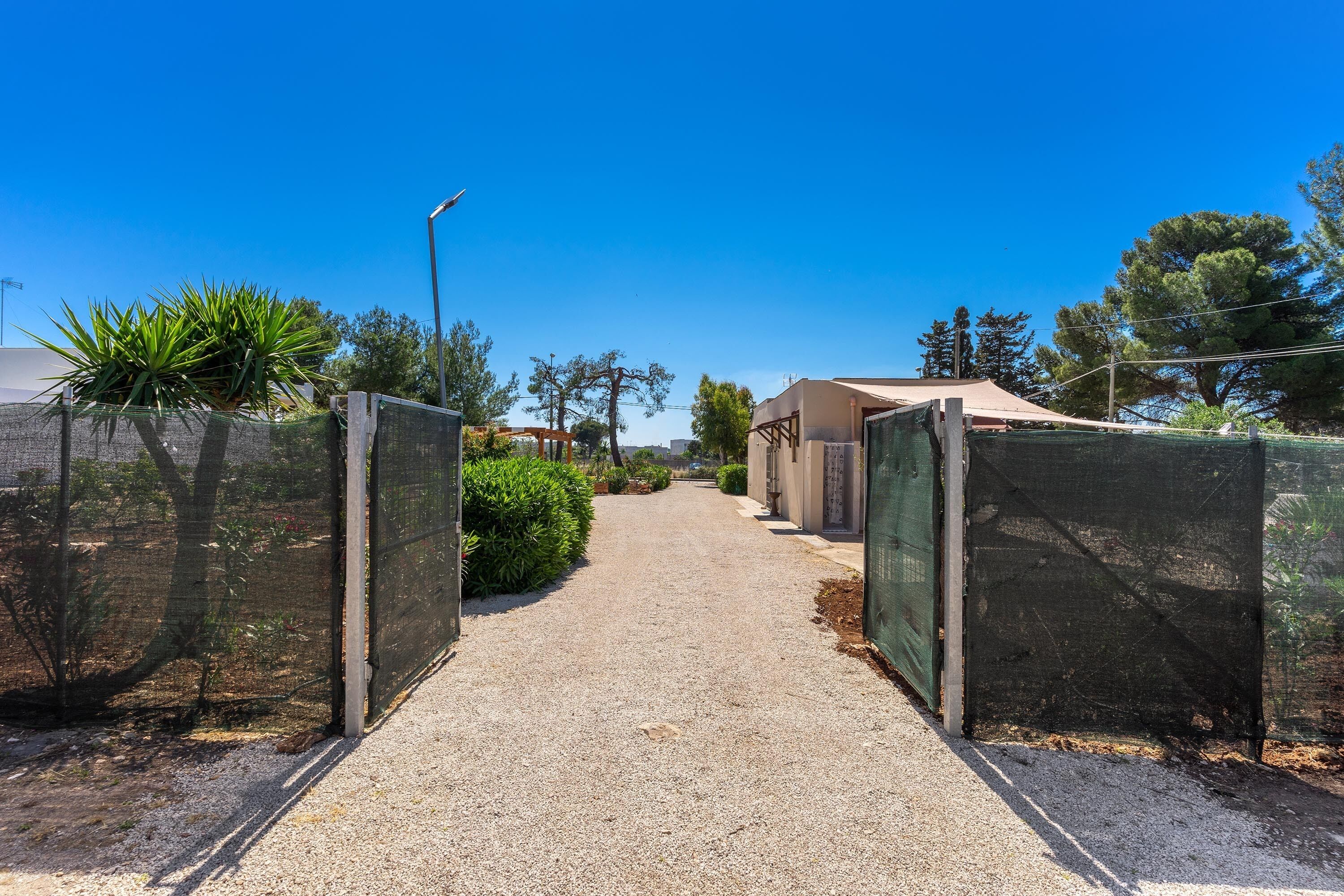 A gravel pathway leading to a building with a covered area, surrounded by greenery and a clear blue sky.
