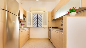 A narrow kitchen with wooden cabinets, a stainless steel refrigerator, a microwave, and a window with a chevron pattern. - 2100 Villetta Martina (Porto Cesareo)