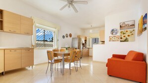 A modern kitchen with wooden cabinets, a dining table with chairs, a refrigerator, and a ceiling fan. - 2100 Villetta Martina (Porto Cesareo)