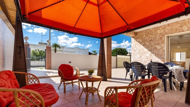 An outdoor patio with wicker furniture, a red umbrella, and a view of the street. - 2100 Villetta Martina (Porto Cesareo)