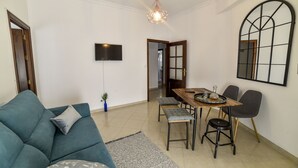 A modern living room with a teal sofa, a dining table with chairs, a TV mounted on the wall, and a large mirror. - Alohamundi San Leandro (Sevilla)