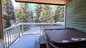 Un'area esterna riparata con un pavimento in legno, una vasca idromassaggio e vista su alberi innevati.