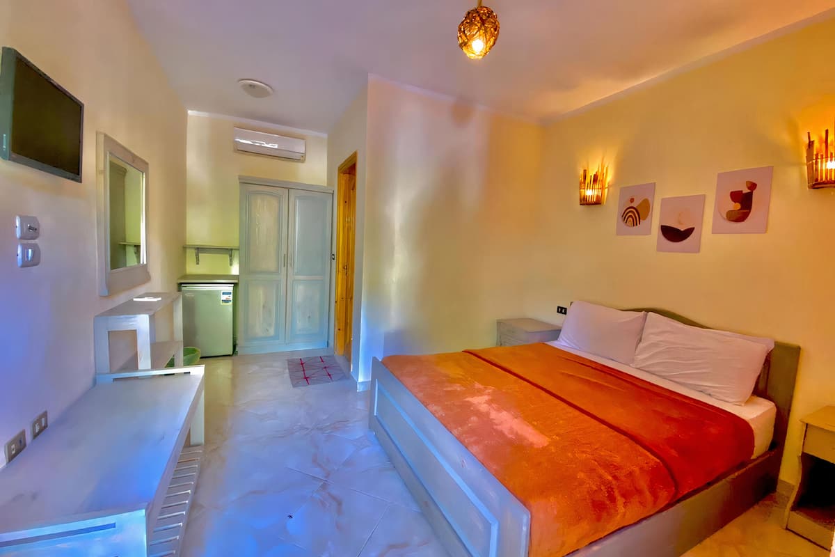 A hotel room with a bed, bedside table, and a desk. There is a kitchenette in the background.