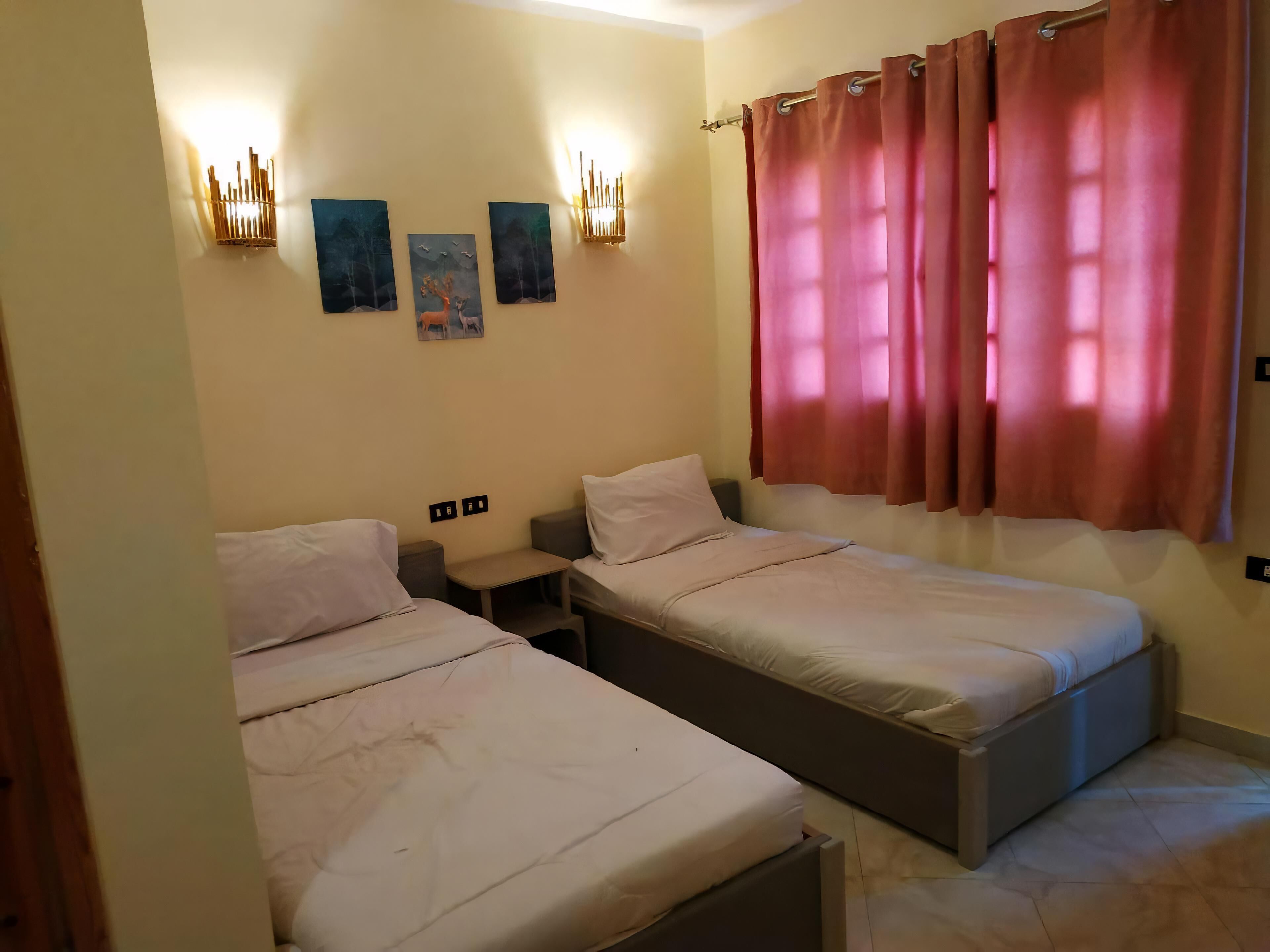 Una habitación con dos camas, una mesita de noche, una lámpara fijada a la pared y un ventanal con cortinas.