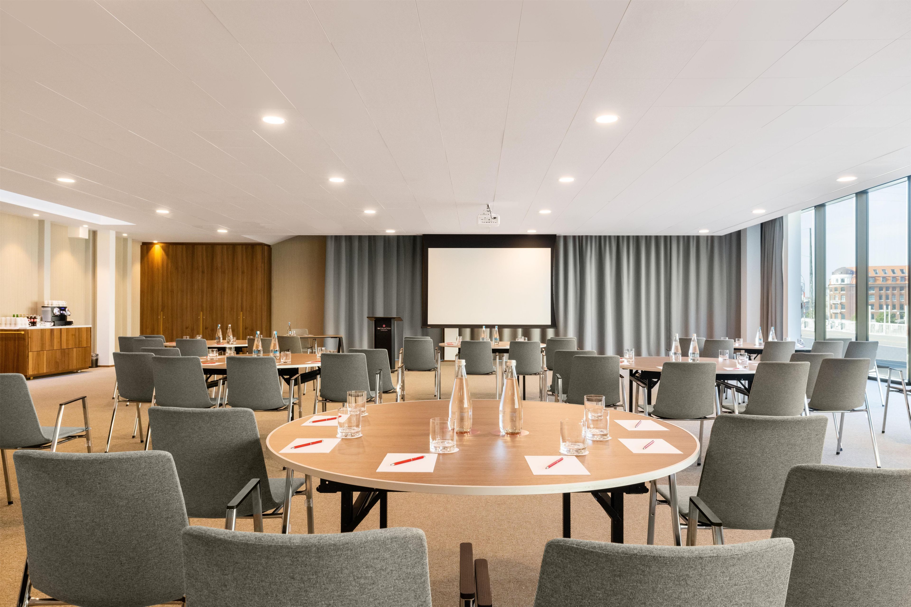 Un salón de conferencias con mesas redondas, sillas grises, un atril y una pantalla de proyector.