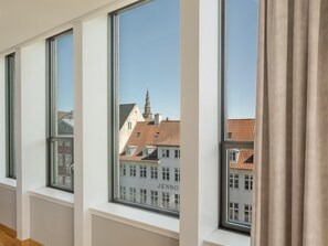 Una habitación con ventanas amplias, paredes blancas y un edificio a la vista con el nombre 'JENNO' en el fondo.