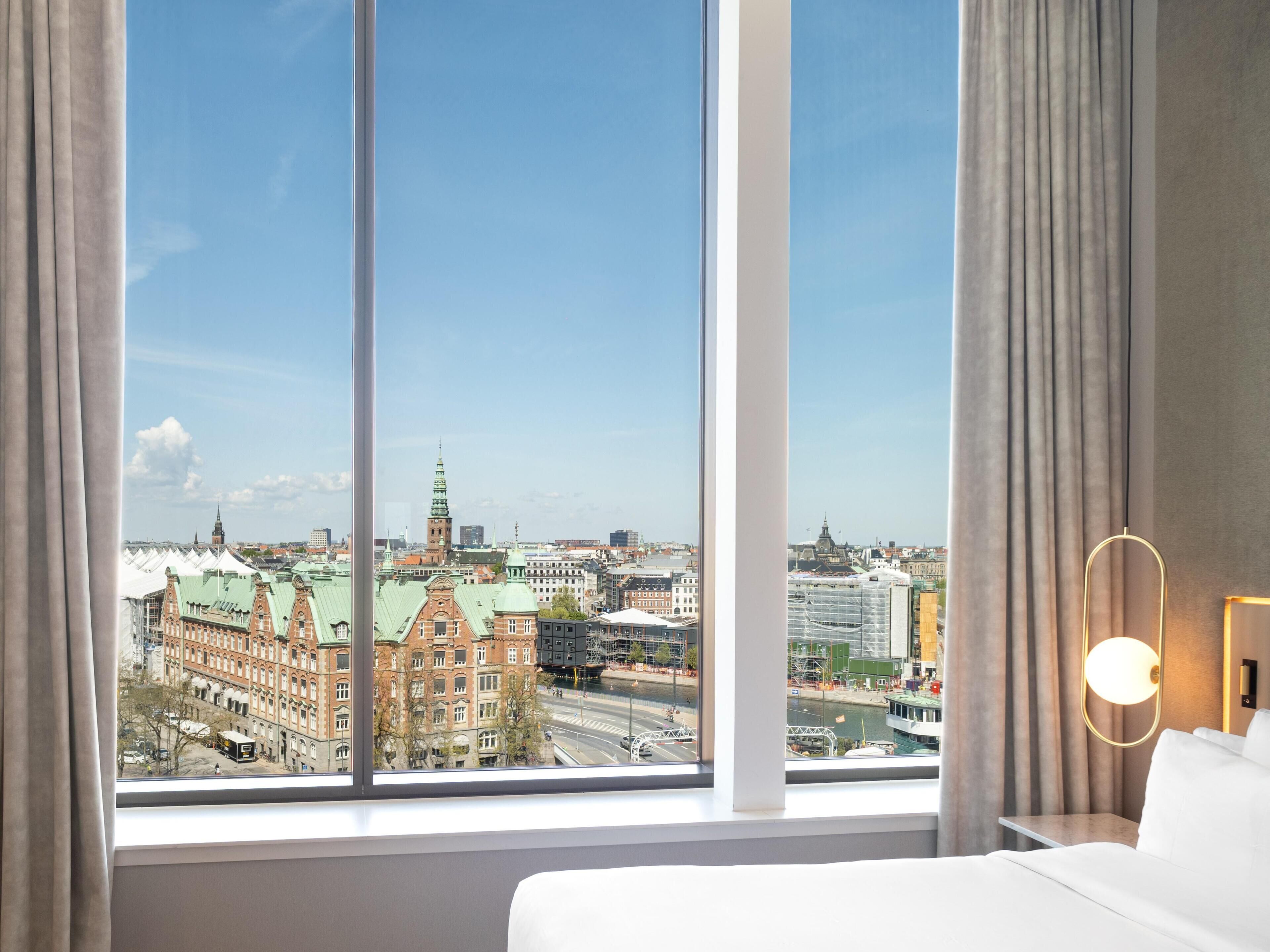 Et hotelværelse med et stort vindue, en seng, et natbord og en lampe.