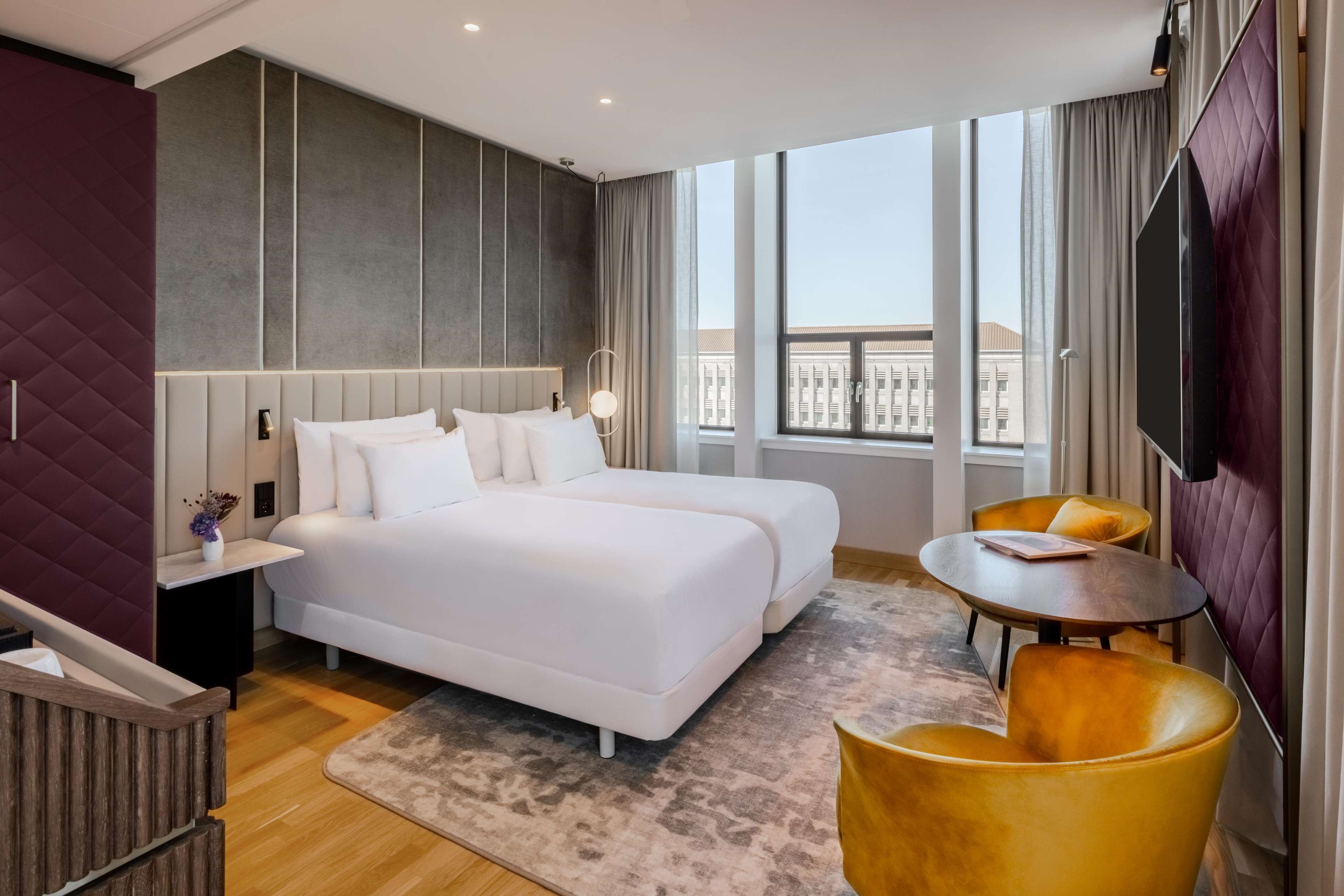 Una habitación de hotel moderna con una cama grande, dos mesitas de noche, una mesita redonda pequeña y un televisor.