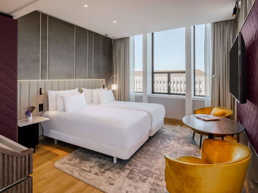 Una habitación de hotel moderna con una cama grande, dos mesitas de noche, una mesita redonda pequeña y un televisor.