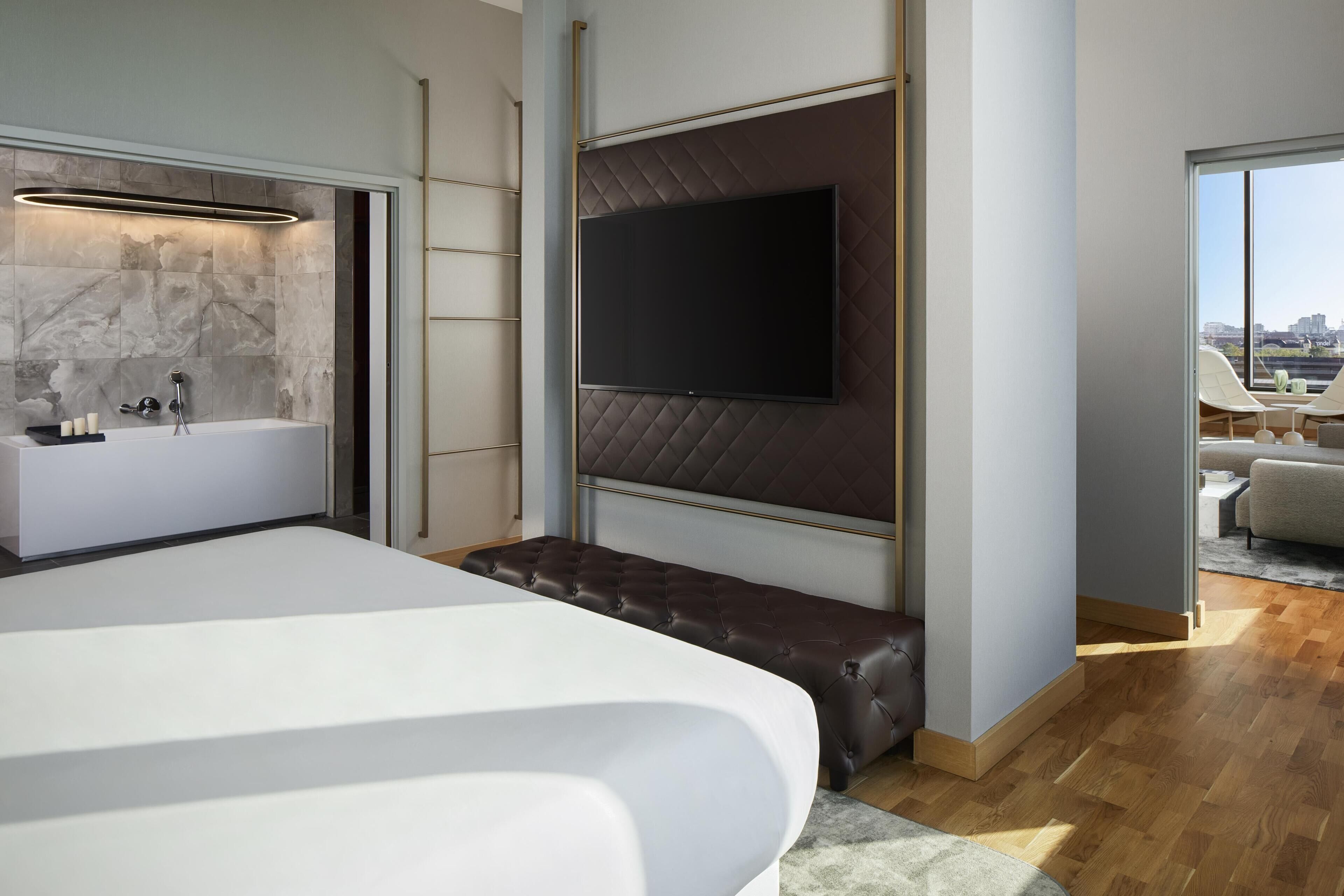 Un dormitorio moderno con una cama grande, una bañera integrada, un televisor de pantalla plana en la pared y vistas de la ciudad a través de un ventanal amplio.