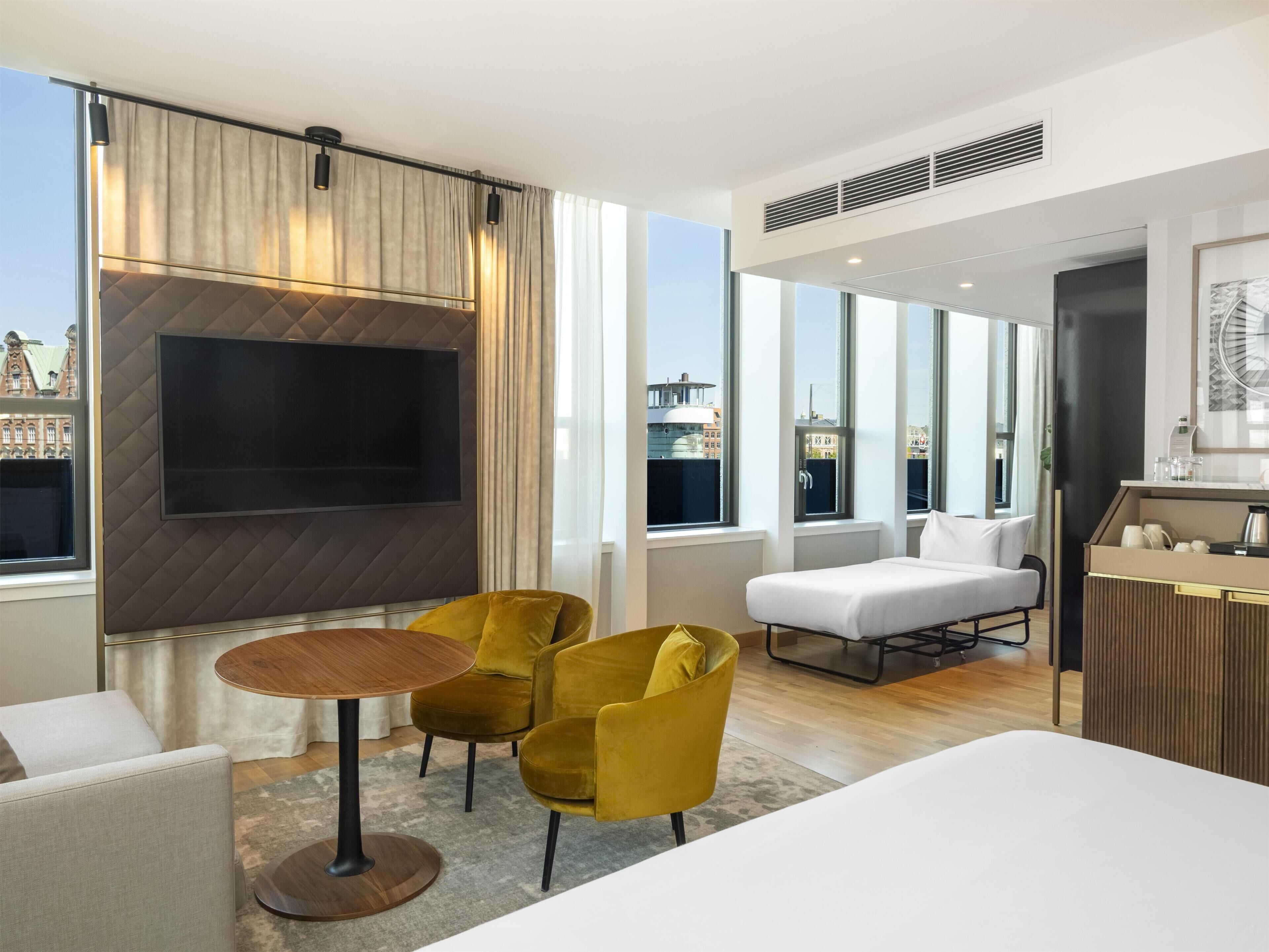 Et moderne hotelværelse med stort fjernsyn, gule stole, en sofa og udsigt over byen.