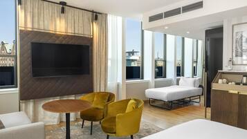 Et moderne hotelværelse med stort fjernsyn, gule stole, en sofa og udsigt over byen.