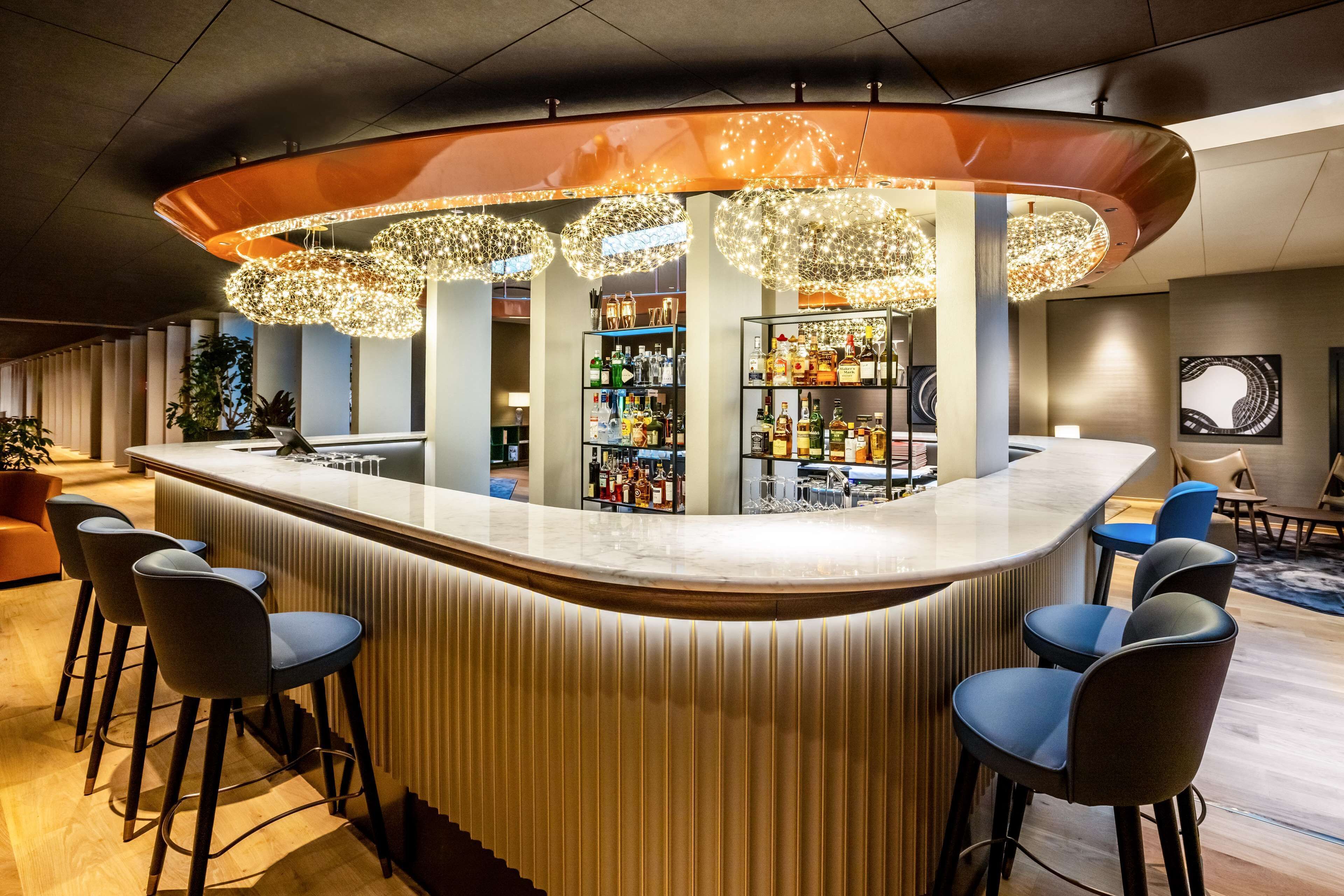 Una zona de bar moderna con una barra curva, taburetes y luces decorativas colgantes.