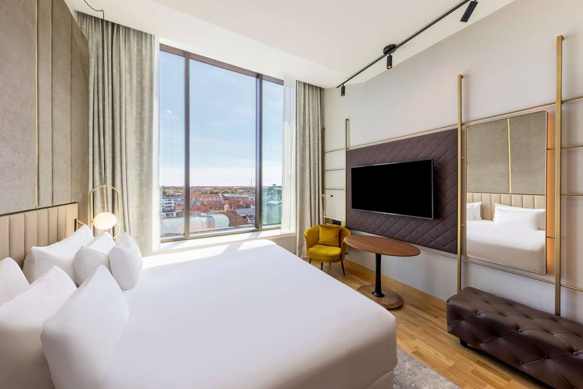 Un moderno cuarto de hotel con una cama grande, un televisor montado en la pared, un sillón amarillo y vistas de la ciudad.