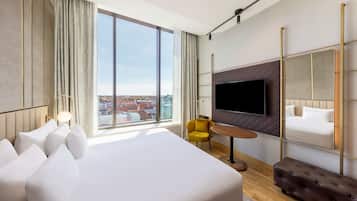 A modern hotel room with a large bed, a TV mounted on the wall, a yellow armchair, and a view of the city.