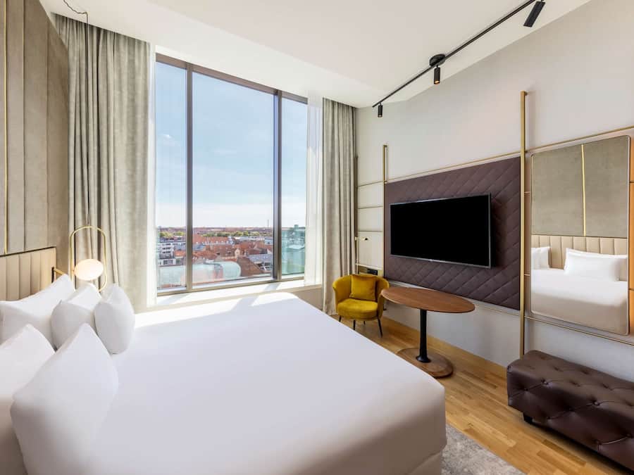 Un moderno cuarto de hotel con una cama grande, un televisor montado en la pared, un sillón amarillo y vistas de la ciudad.
