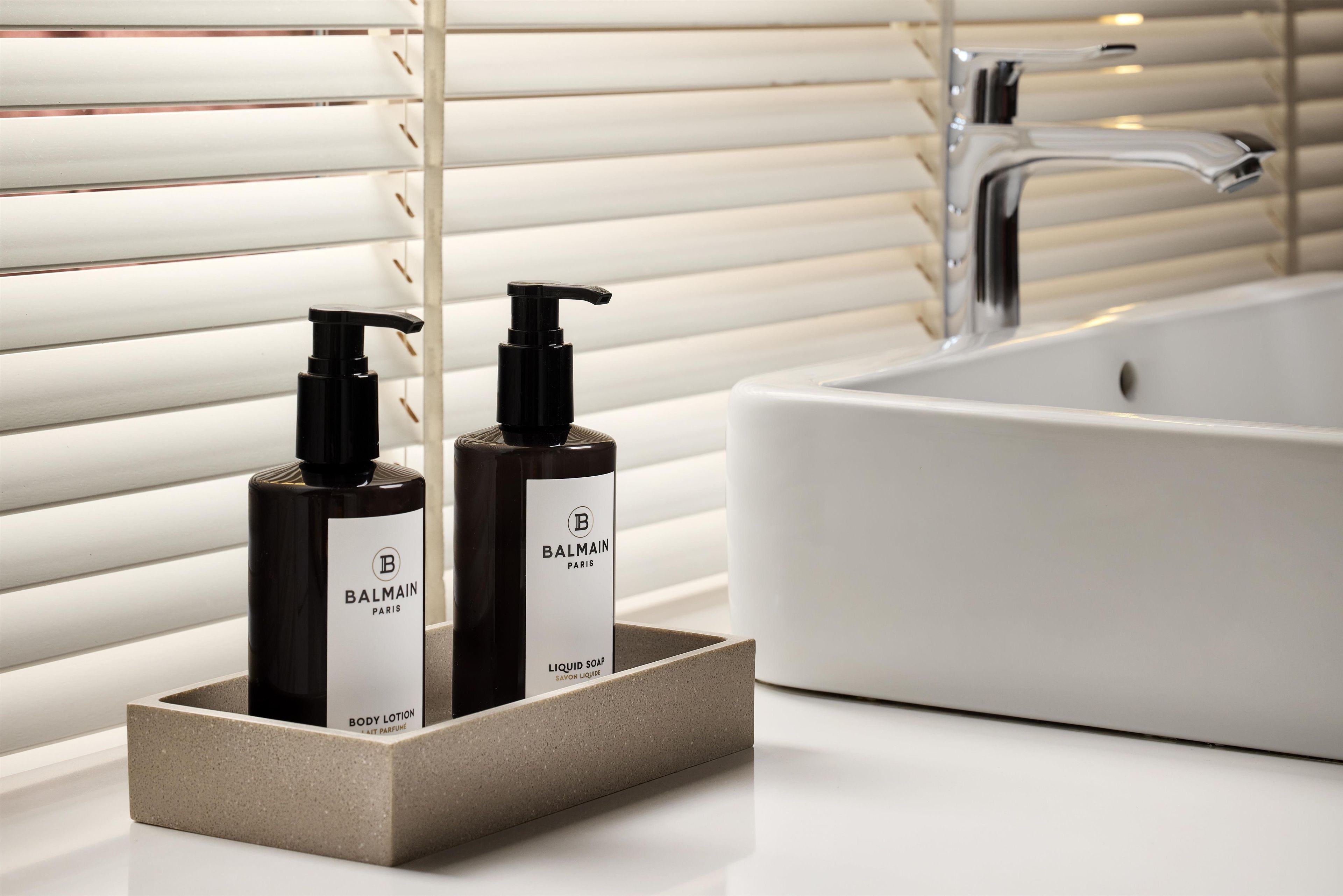 Une salle de bain avec un lavabo blanc, deux flacons de lotion pour le corps de la marque Balmain et un robinet moderne.