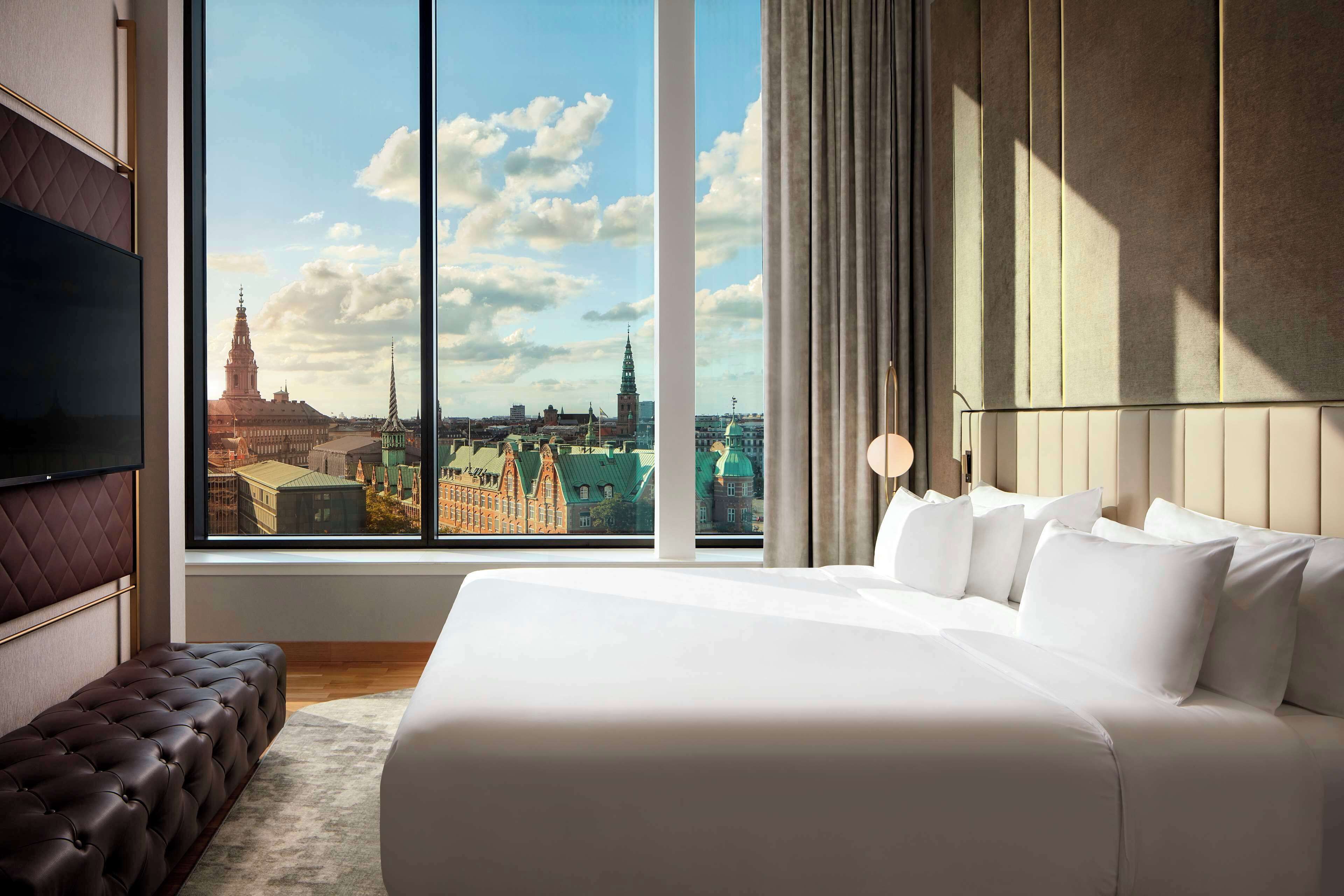 A modern hotel room with a large window offering a cityscape view, a plush bed, and a seating area.