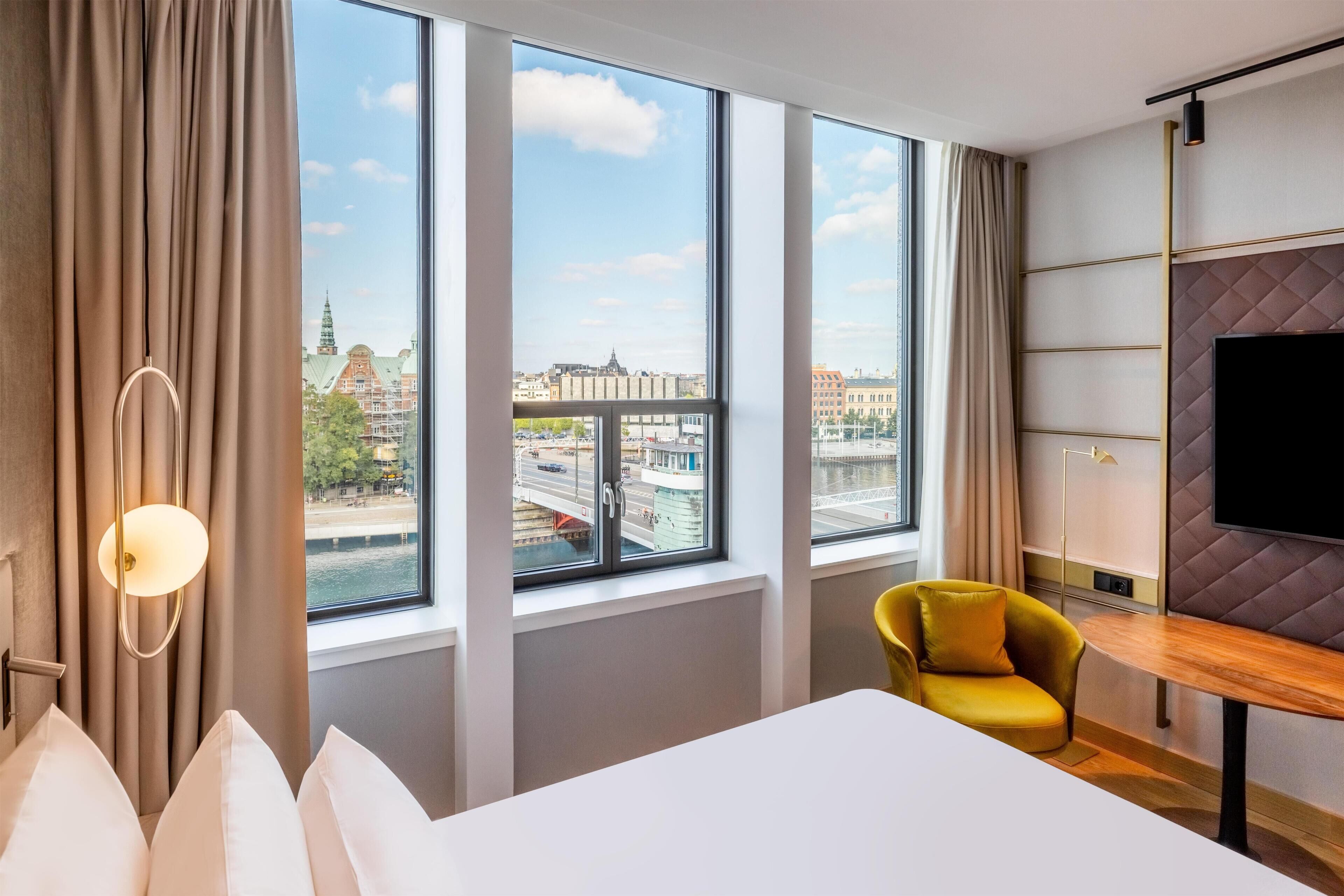 A hotel room with a large window, a bed, a yellow armchair, and a flat-screen TV.