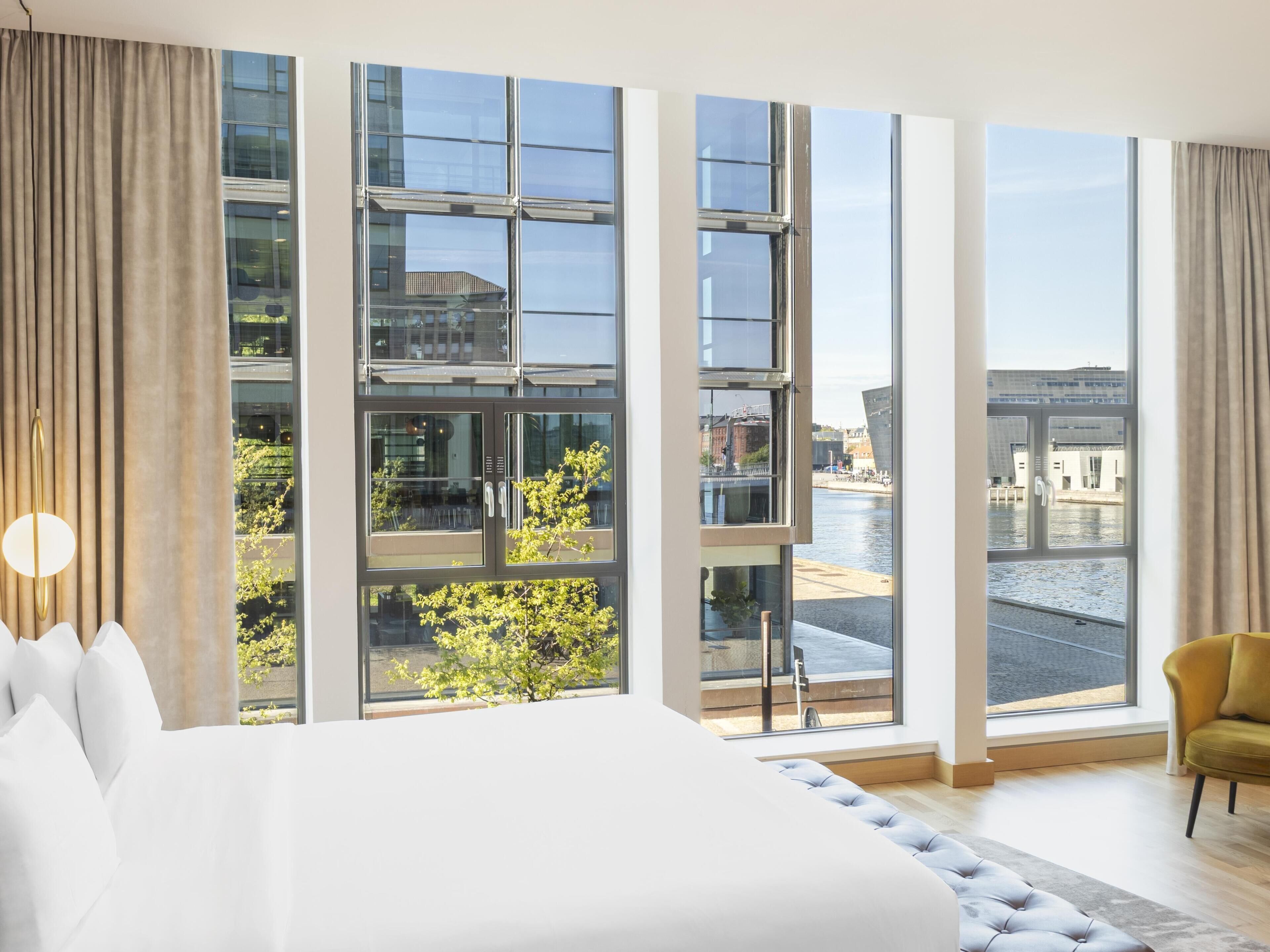 A modern hotel room with large windows, a bed, a seating area, and a view of the city.
