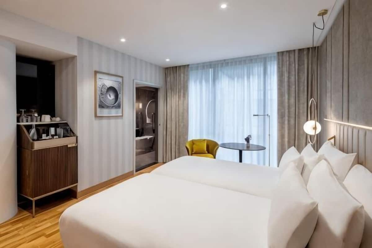 Habitación de hotel con una cama grande, una mesita, una silla amarilla, un tocador con artículos de aseo y un cuadro en la pared.
