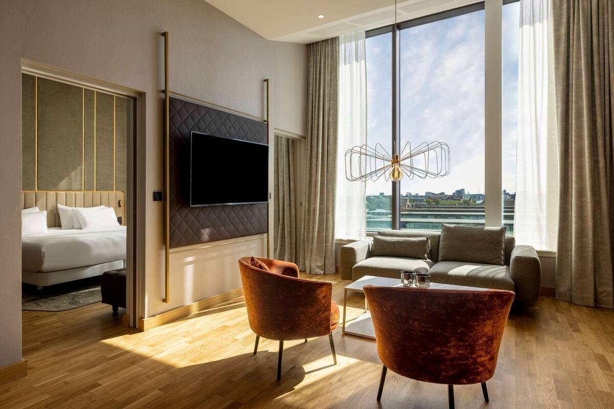 Una habitación de hotel moderna con una cama grande, un televisor montado en la pared, un sofá, dos sillones, una mesita de centro y vistas a la ciudad a través de amplios ventanales.