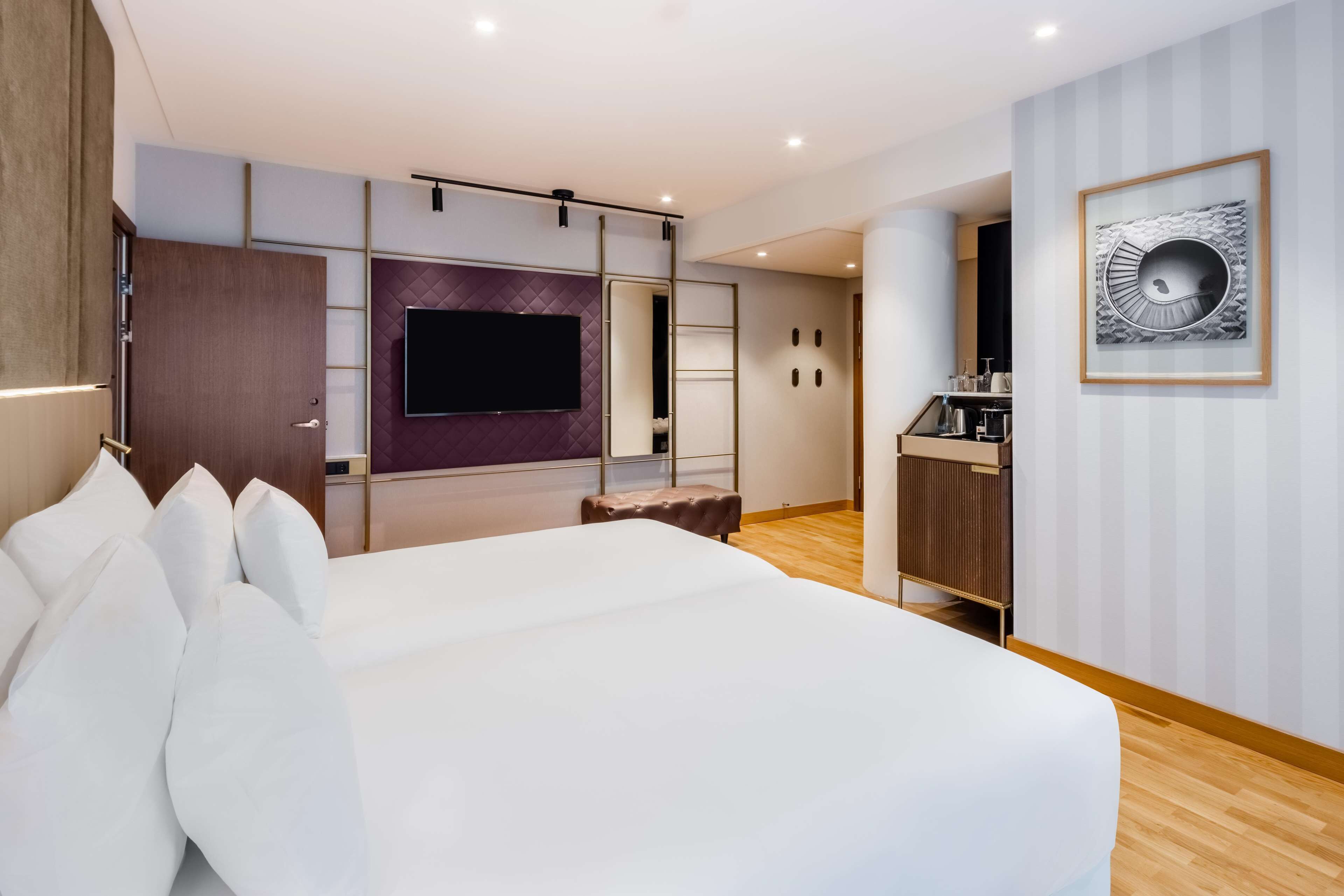 A modern hotel room with a large bed, a flat-screen TV mounted on the wall, a small seating area, and a kitchenette.