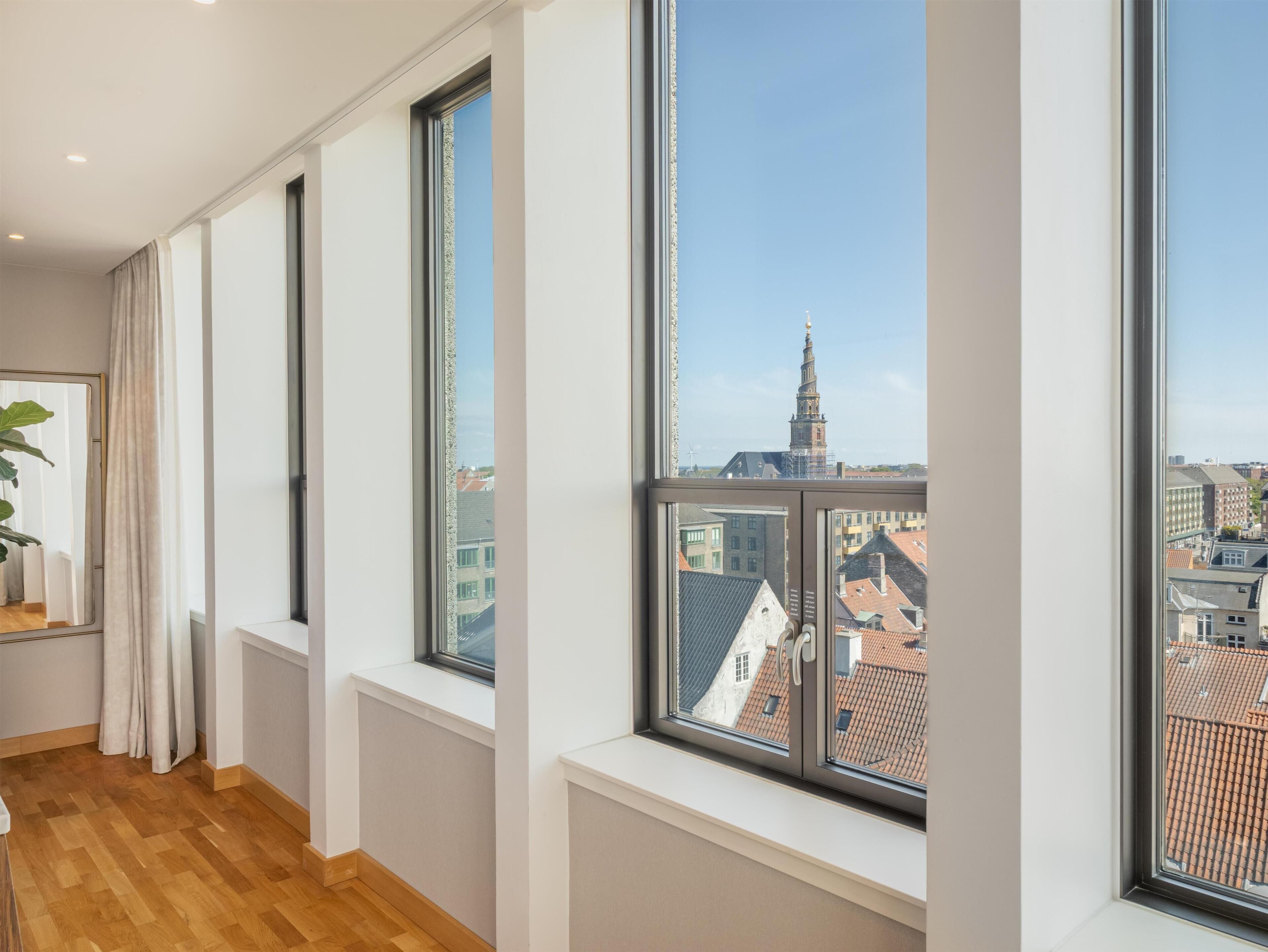 Una habitación con ventanas amplias, piso de madera y vistas a edificios y una torre.