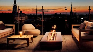 Terraza en la azotea con una chimenea integrada de diseño moderno, asientos cómodos y vista al horizonte de la ciudad al atardecer.