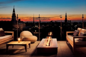 A rooftop terrace with a modern fire pit table, comfortable seating, and a city skyline view at dusk. - NH Collection Copenhagen (Copenhagen)