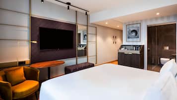 Una habitación de hotel moderna con una cama grande, una zona de estar con una silla y una mesa, un televisor de pantalla plana en la pared, una kitchenette pequeña y un baño visible a través de una puerta de cristal.