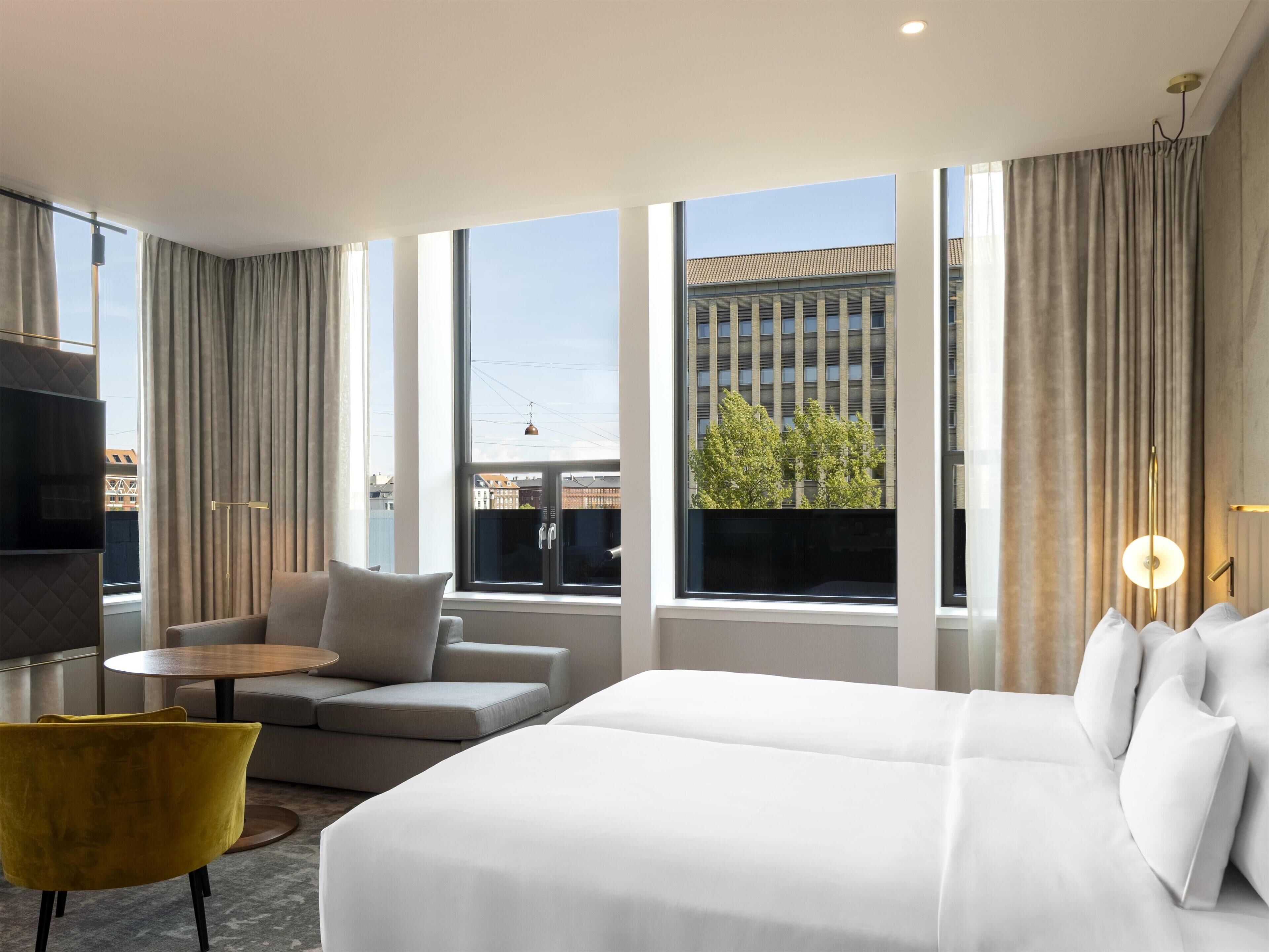 Habitación de hotel con cama, sofá, una mesita, una silla amarilla y ventanas grandes con cortinas.