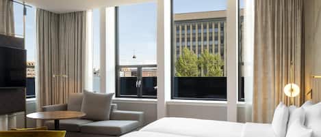 A hotel room with a bed, a sofa, a small table, a yellow chair, and large windows with curtains.