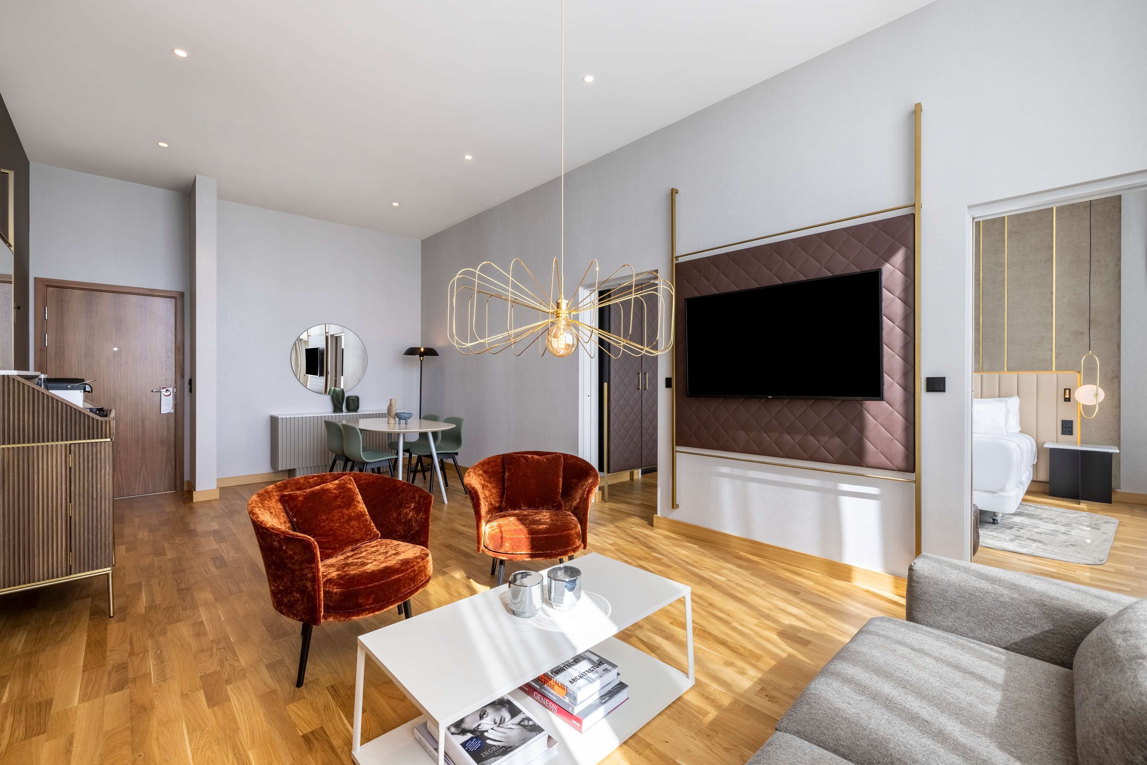 A modern living room with a flat-screen TV, velvet chairs, a coffee table with magazines, and a unique ceiling light.