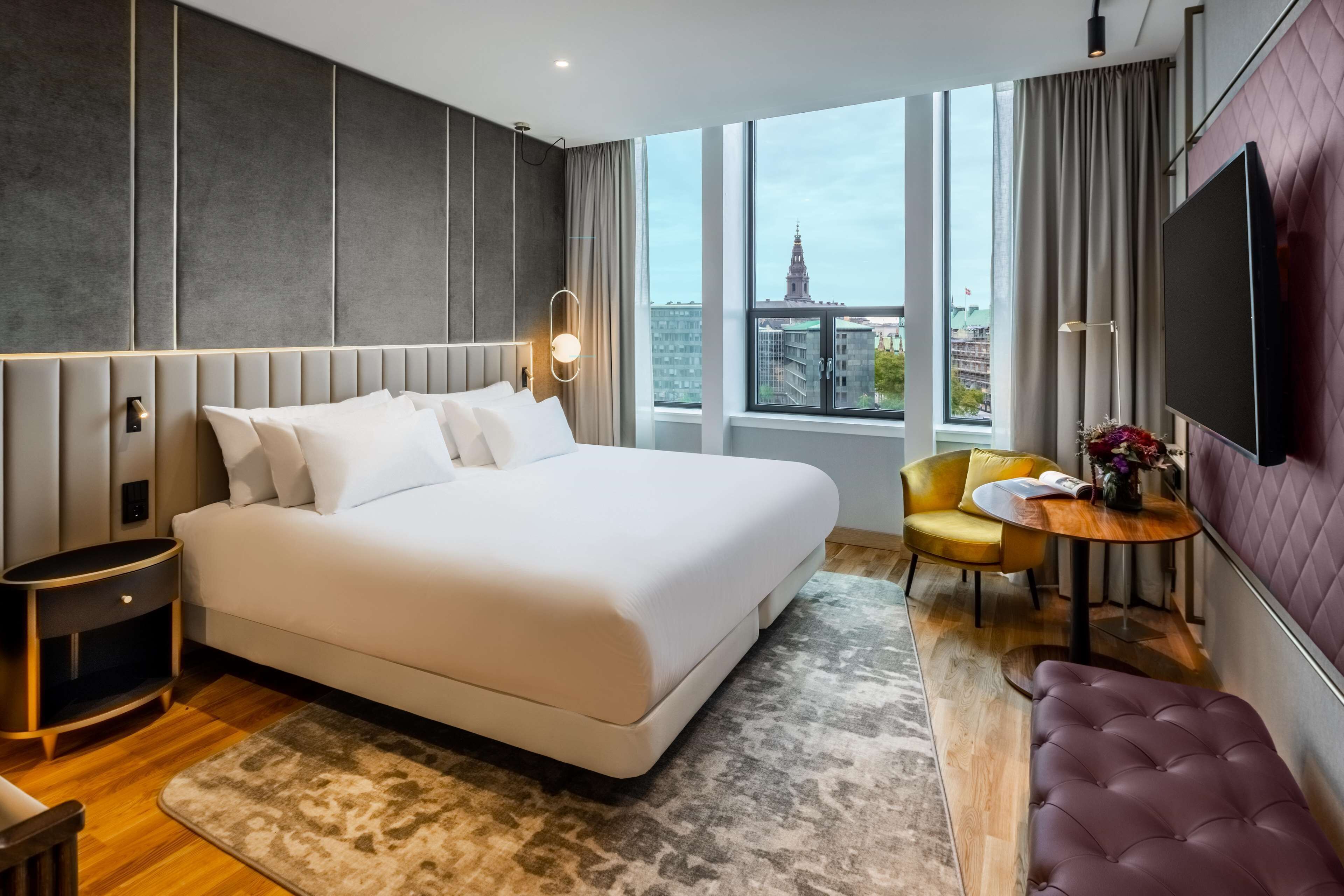 A modern hotel room with a large bed, a bedside table, a flat-screen TV, a small round table with a vase of flowers, a yellow chair, and a purple bench.