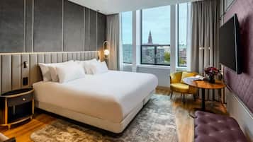 Ein modernes Hotelzimmer mit einem großen Bett, einem Nachttisch, einem Flachbildfernseher, einem kleinen runden Tisch mit Blumen und einem gelben Sessel sowie einer violetten Bank.